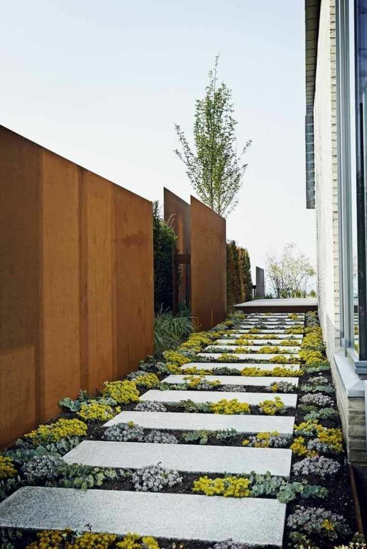Sekretessskärm av cortenstål av enkla paneler längs en trädgårdsväg av stenplattor