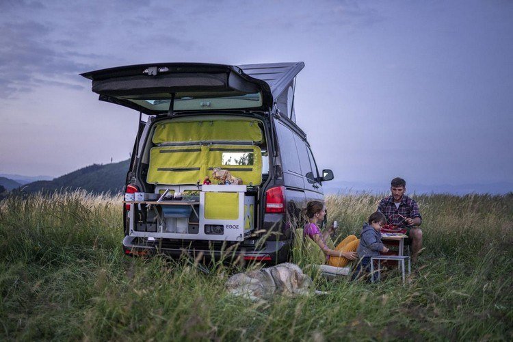 Familj med barn som äter middag i naturen med ett campingfordon utrustat med en campingbox