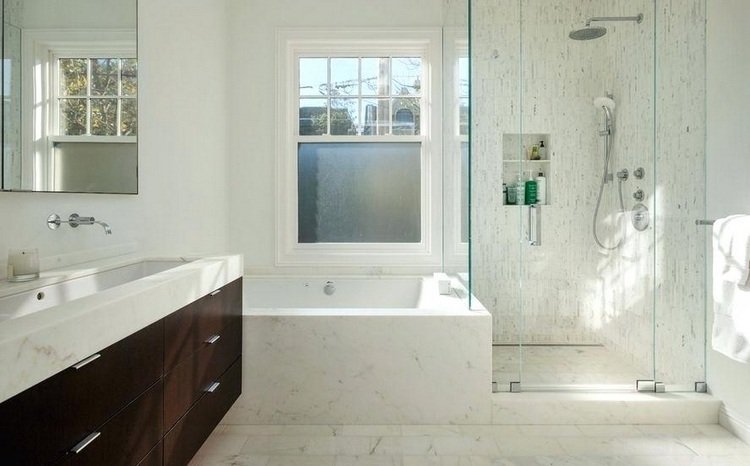 Badkar framför fönstret med duschkabin och nisch i badrummet i vit marmor och trä