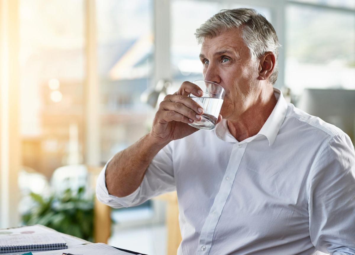 Äldre man med grått hår dricker från vattenglas för bättre vattenabsorption för att bli av med ölmage