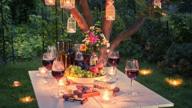 Matbord med vacker trädgårdsfestbelysning - ljus på bordet, i gräsmattan och hängande på trädet i fågelburar