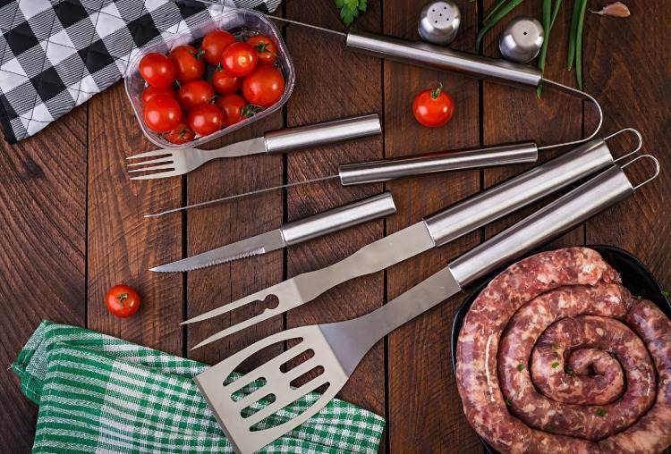 grilltillbehör som spateltång och vass kniv på träbord med korv och tomater