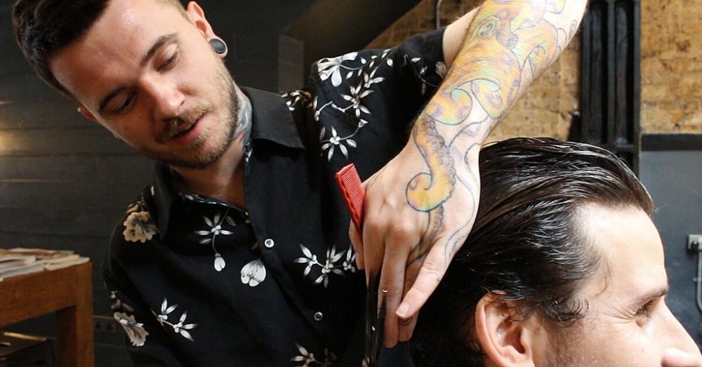Frisör med tatuering på armen kammar mans långa hår i frisörbutiken