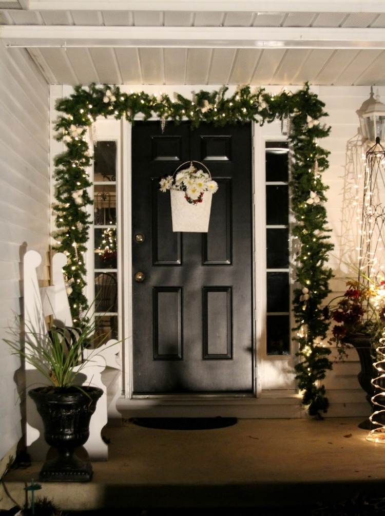 Juldekoration-utanför-upplyst-ytterdörr-grangrenar-dekorerade-fe-lampor-svart-vit-entré