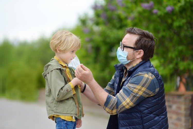 Far med skyddsmask sätter munskydd på sin sons ansikte under covid 19 -pandemin
