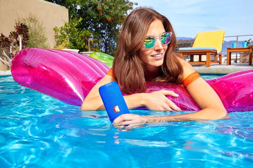 Kvinnan i solglasögon ligger under solen i poolen på vattenmadrass och håller trådlös högtalare