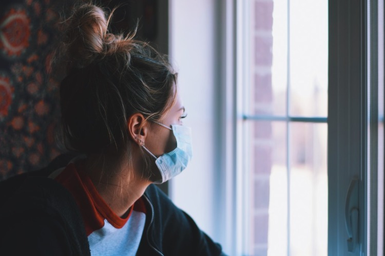 Ung kvinna med skyddsmask tittar ut genom fönstret under Covid -19 -pandemin