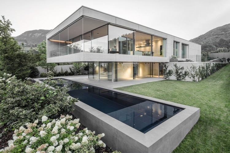 exponerat betonghus casa mf stora glasfönster modern minimalistisk design framifrån gräsmatta pool bergssluttning blommor