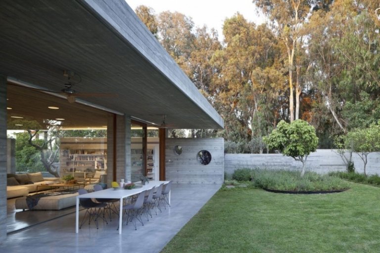 Exponerad betong inuti -trädgård-gräsmatta-matbord-stolar-vardagsrum-glasvägg-träd-buskar