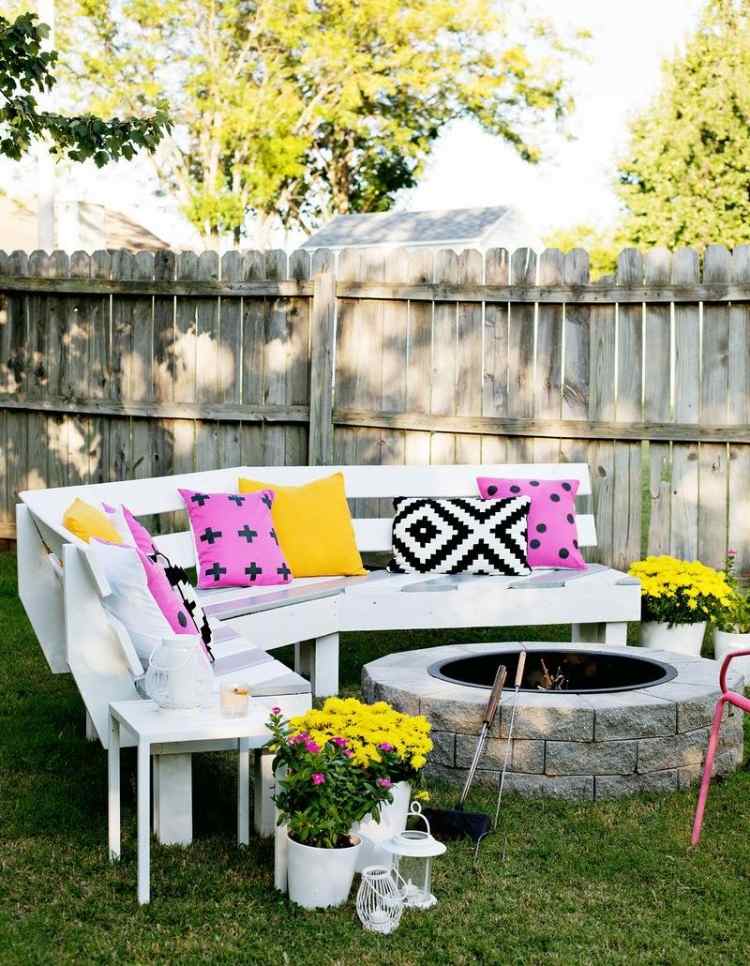 öppen spis-trädgård-sits-bänk-trä-vita-kuddar-färgade-runda-stenar-planter-blommor-rosa-gul