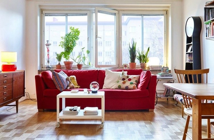 Ställ upp soffa framför fönstret vardagsrum snygg soffa röd uppsatt soffbord bäddsoffa rullkuddar färgglada retro stil väggklocka växter matbord