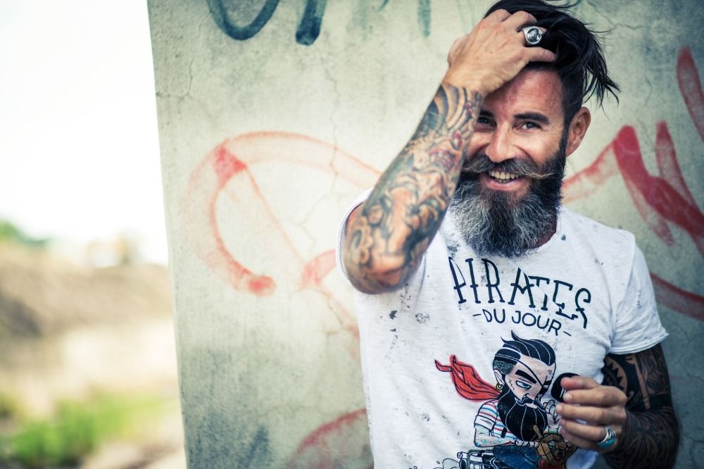 skrattande man med skägg och modern frisyr visar armen tatueringar män
