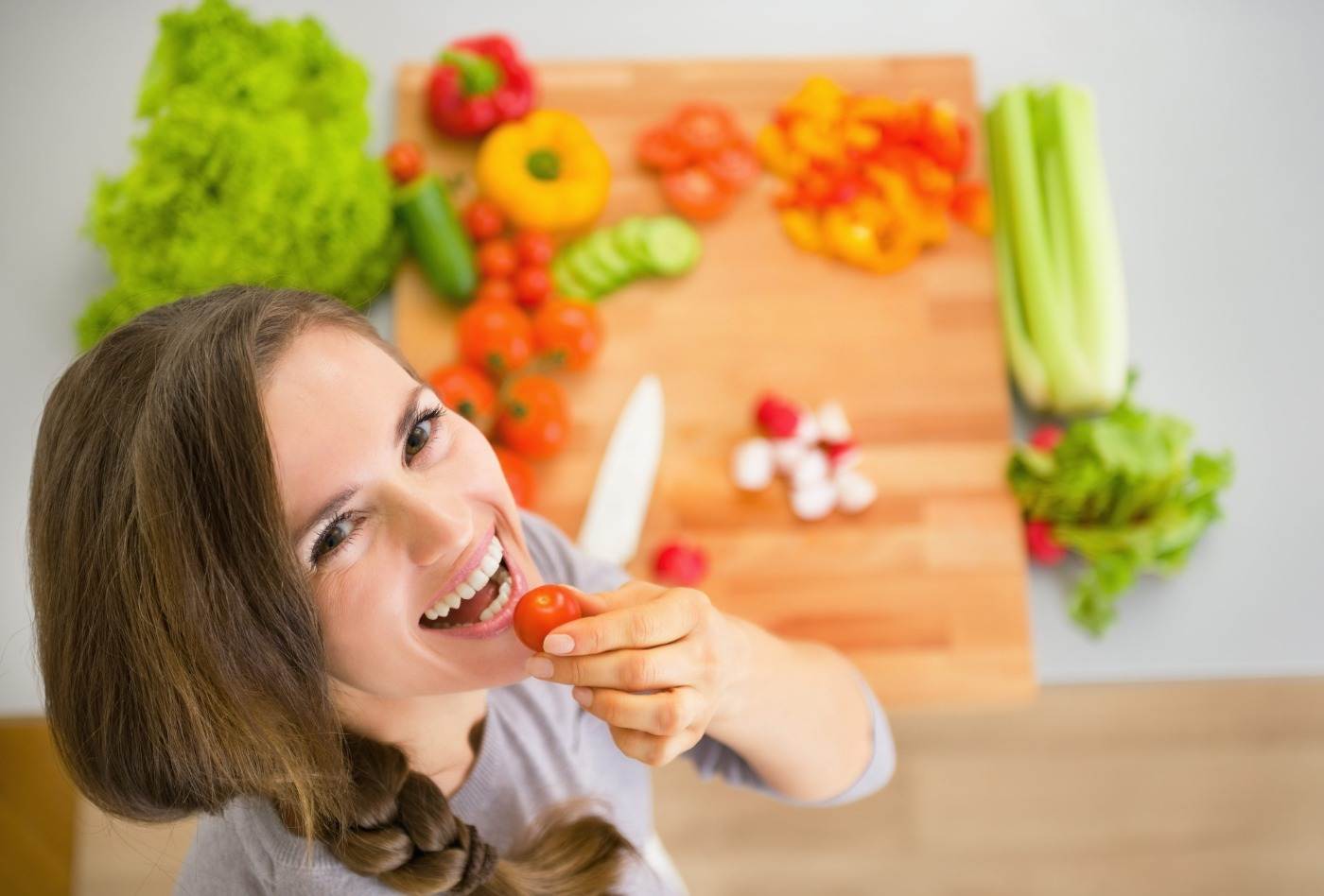 Kvinna som ler och håller körsbärstomat framför munnen med grönsaker och köksbräda i bakgrunden