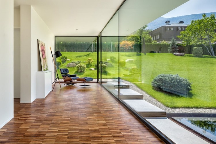 glas-vägg-inuti-utsida-ljus-bambu-golv-panorama-trädgård-fåtölj-vy